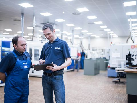 zwei männliche Mitarbeiter von Häring, ein Mitarbeiter trägt ein Blaumann und schaut auf ein Tablet von einem anderen Mitarbeiter in einem blauen T-Shirt unterhalten sich